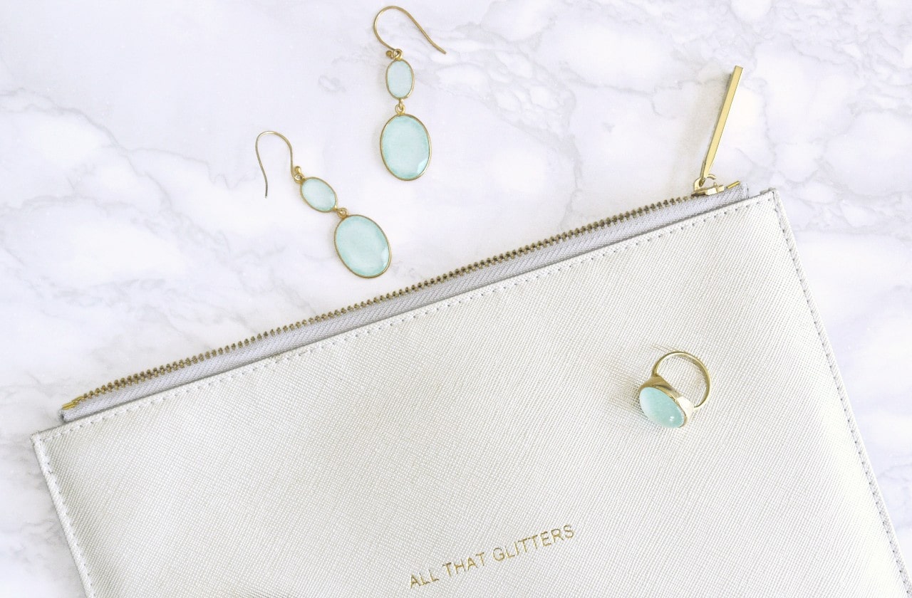 A matching pair of turquoise drop earrings and bezel statement ring lay on a marble bathroom vanity with a white leather makeup bag