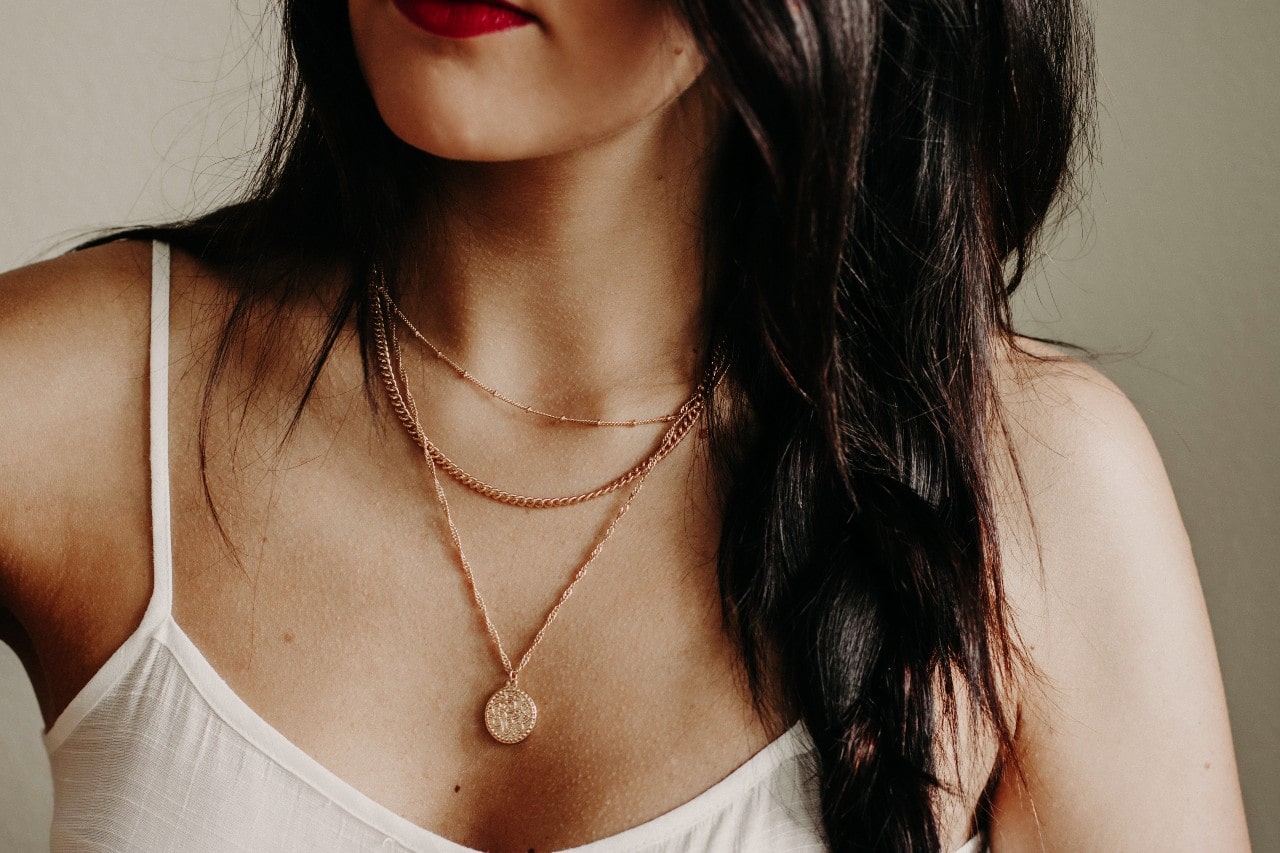 A young woman in a white tank top wears three layered yellow gold necklaces to express her personal style