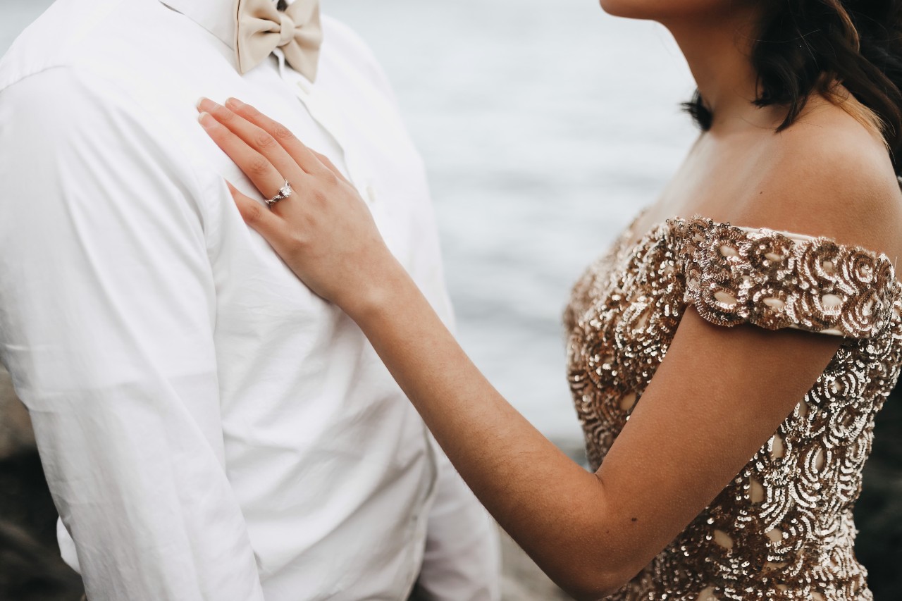 A couple in formal wear; her in a champagne sequin off the shoulder gown and him in a white button down and a matching bow tie