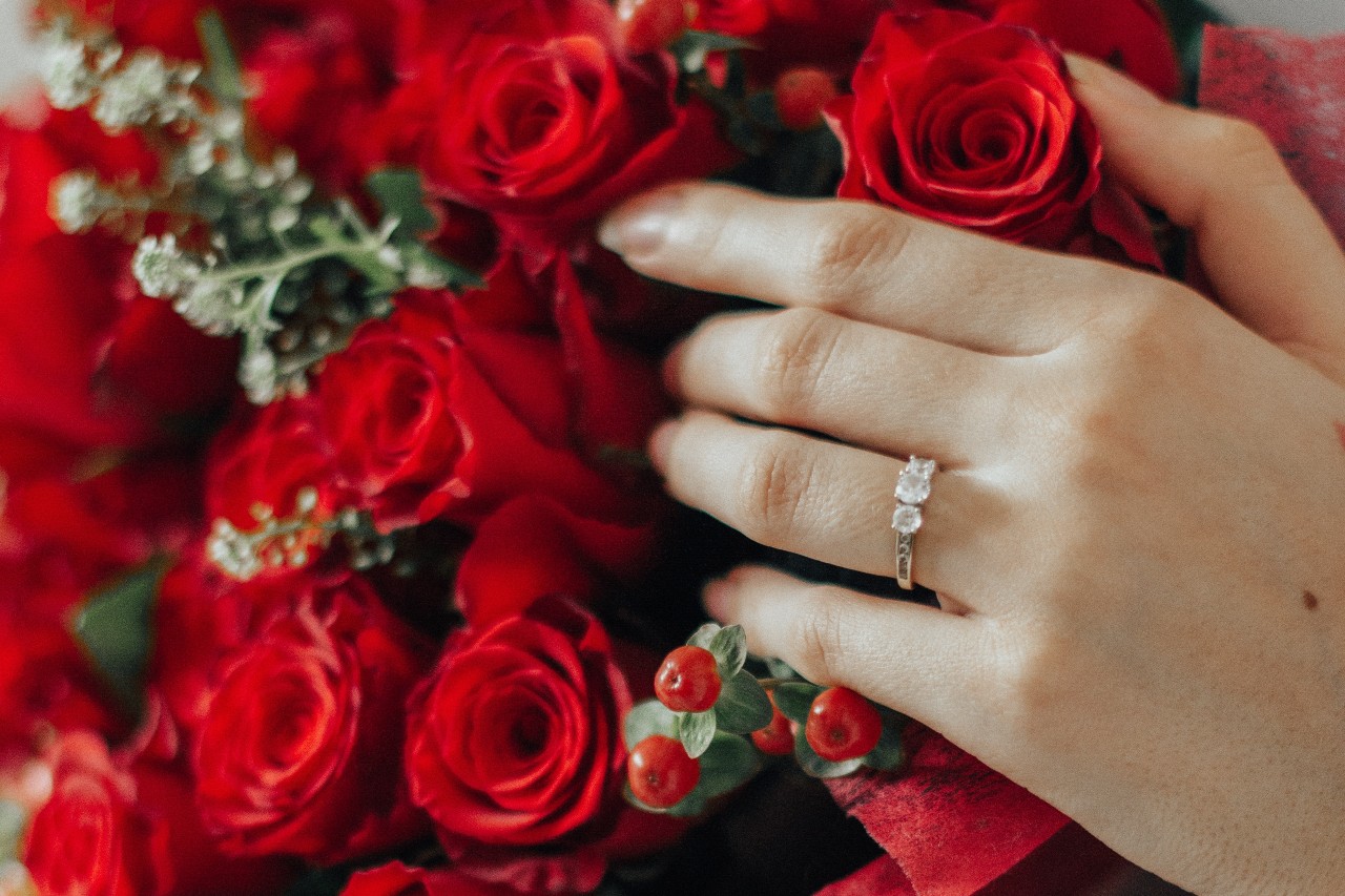 A hand with a three stone engagement ring sitting on top of a bouquet of red roses