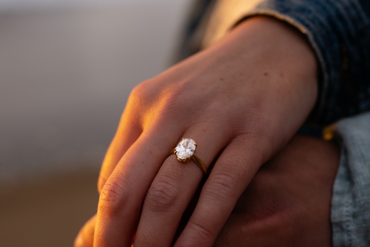 A woman’s hand resting on her partner’s, adorned with an oval cut engagement ring.