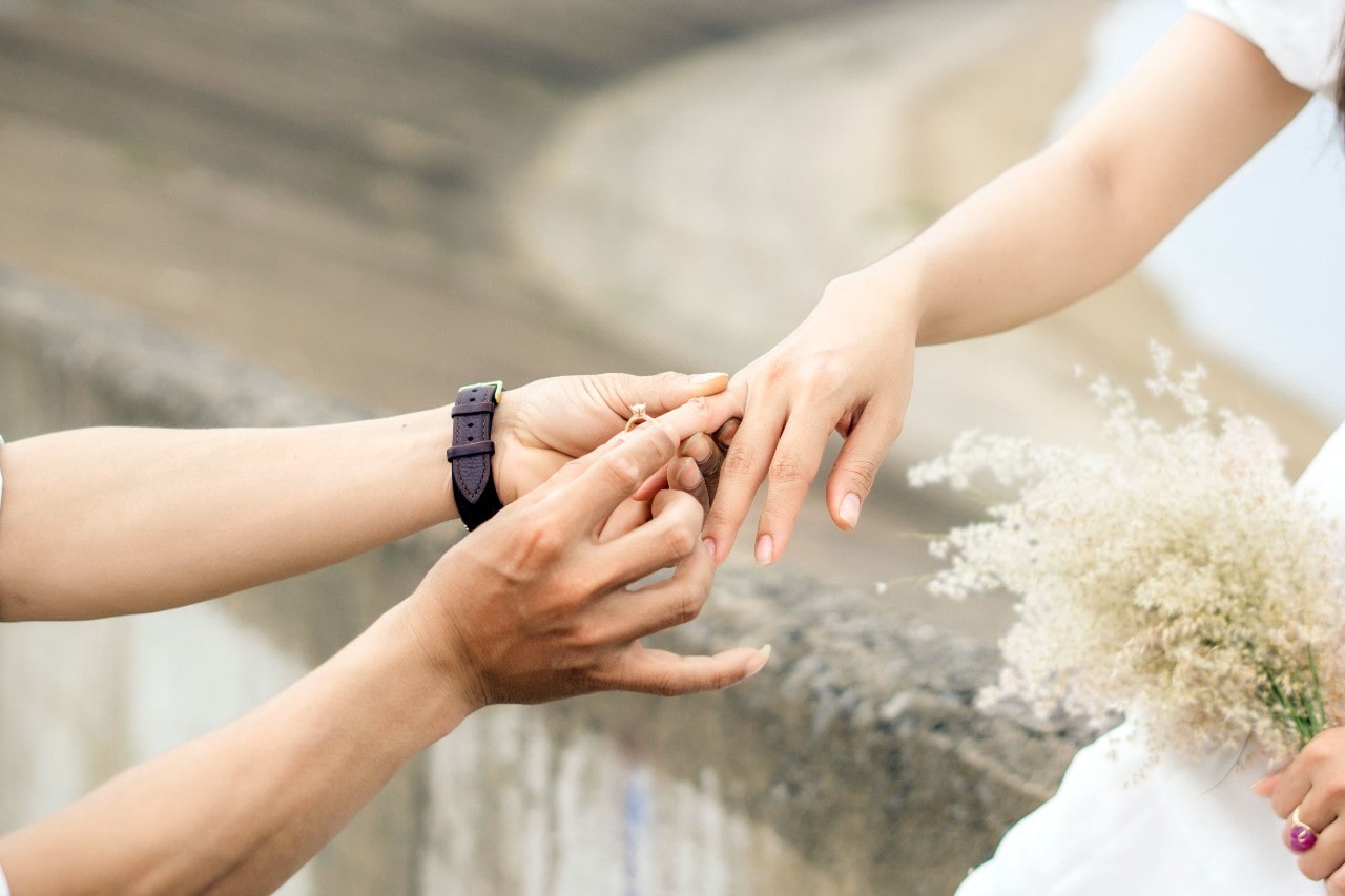 A person placing an engagement ring on their bride-to-be’s finger.