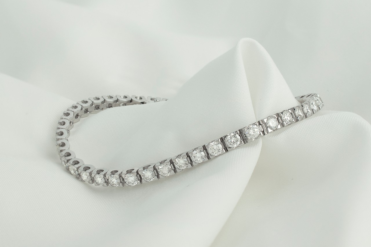 A close-up of a delicate diamond tennis bracelet displayed atop drapey white fabric.