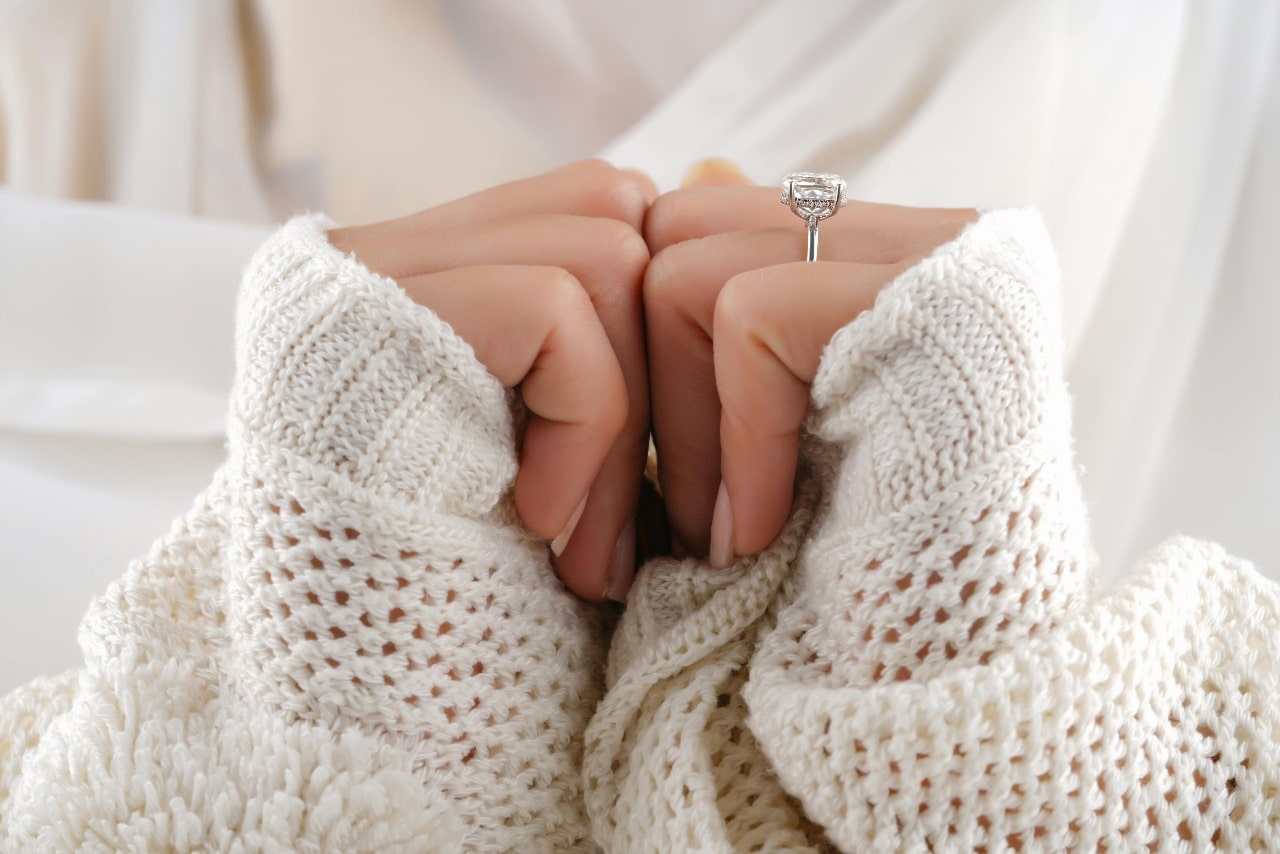 A woman’s hands adorned with a silver engagement ring, wrapped in a white chunky blanket.