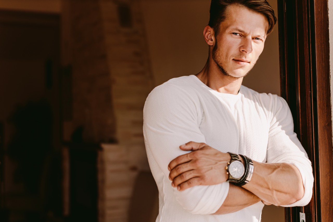 A blonde man in a white shirt leaning casually against a wall, wearing a large wristwatch.