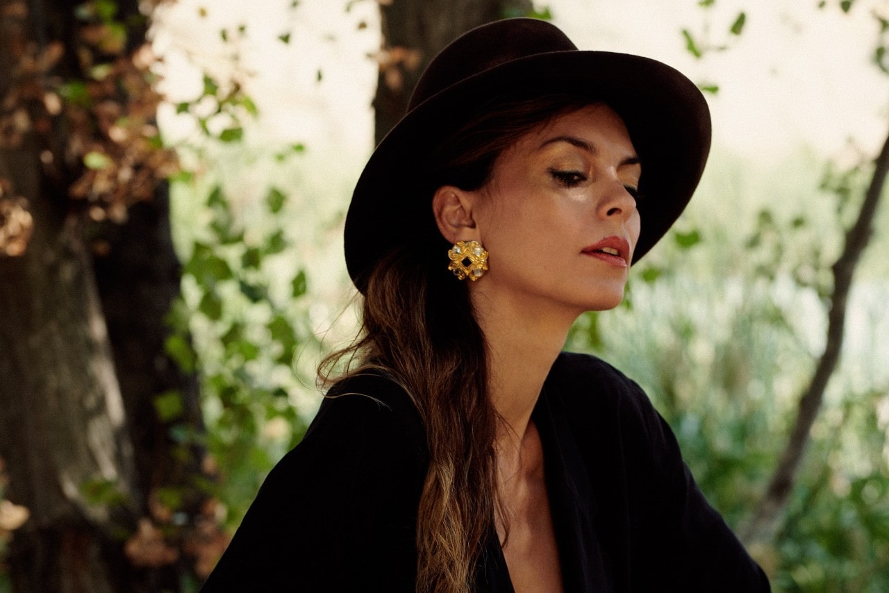 A woman wearing chunky gold stud earrings sits under a tree.