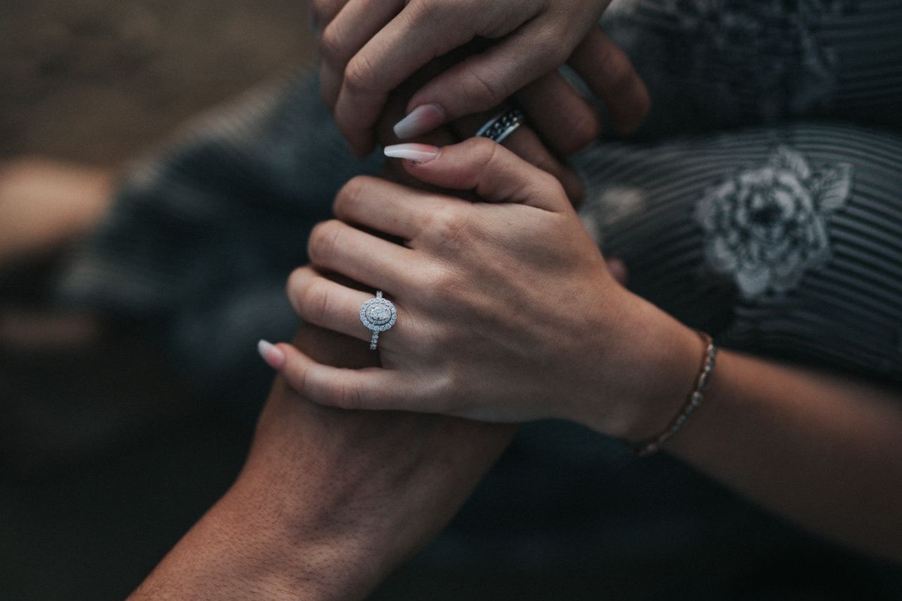 A woman wearing a halo engagement ring holding her fiance’s hands.