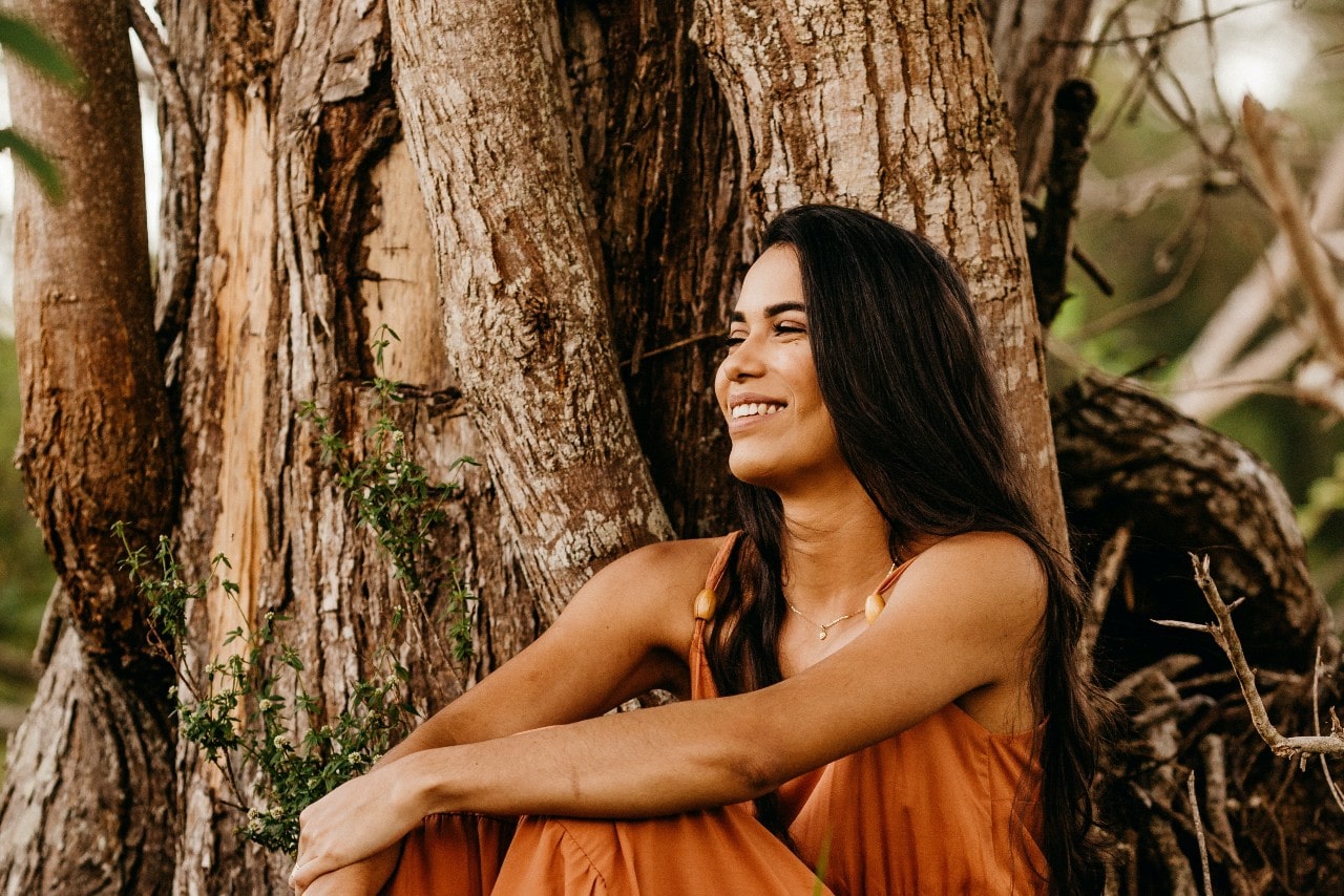 a woman sitting by a tree smiling and wearing a simple necklace