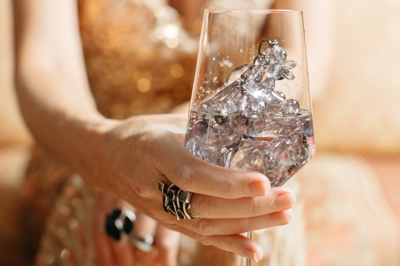 A close-up of an older woman’s hand holding a wine glass, half-full with water and shining stones.