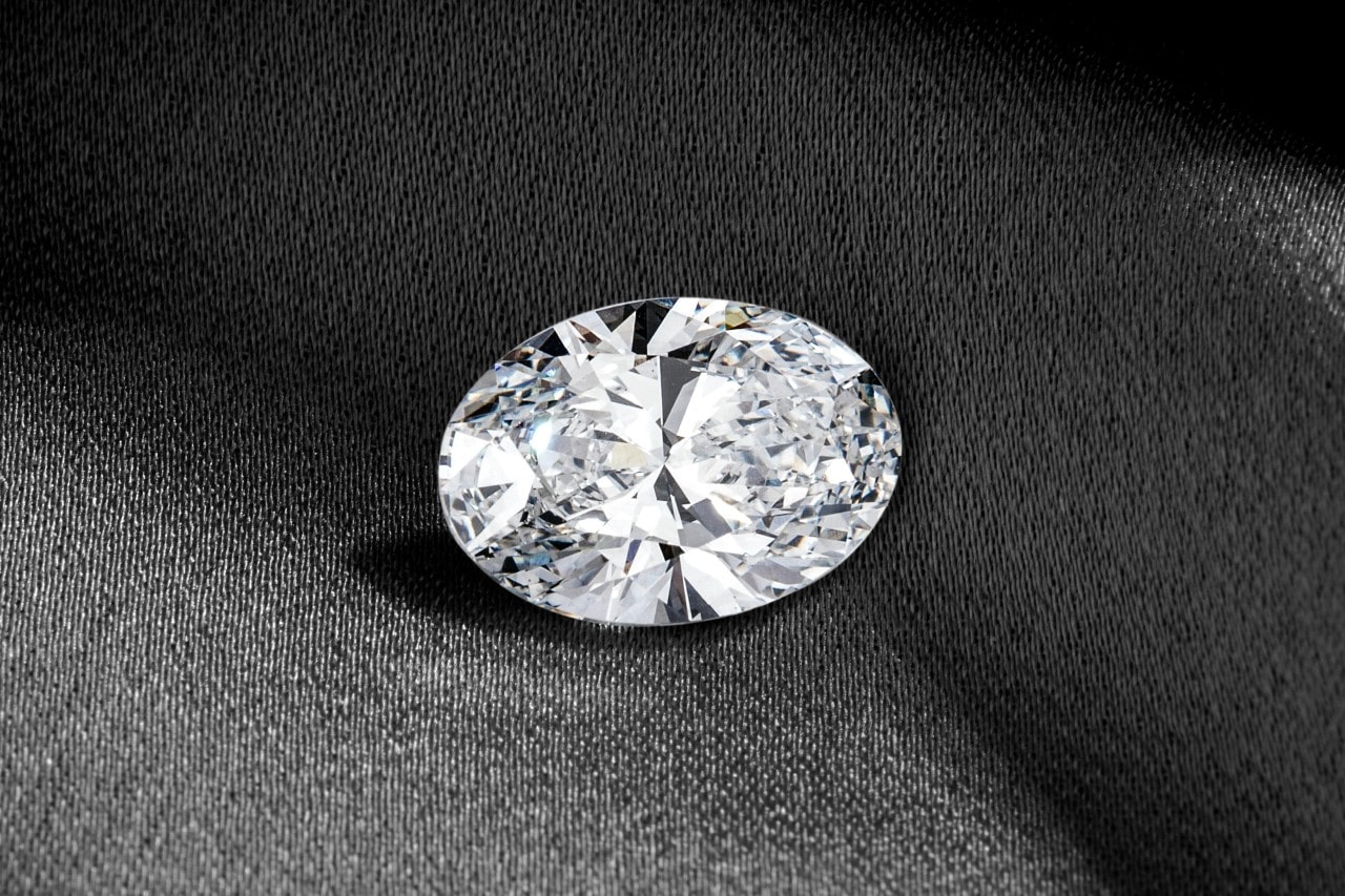 A close-up of a glistening oval-cut diamond on a dark fabric background.