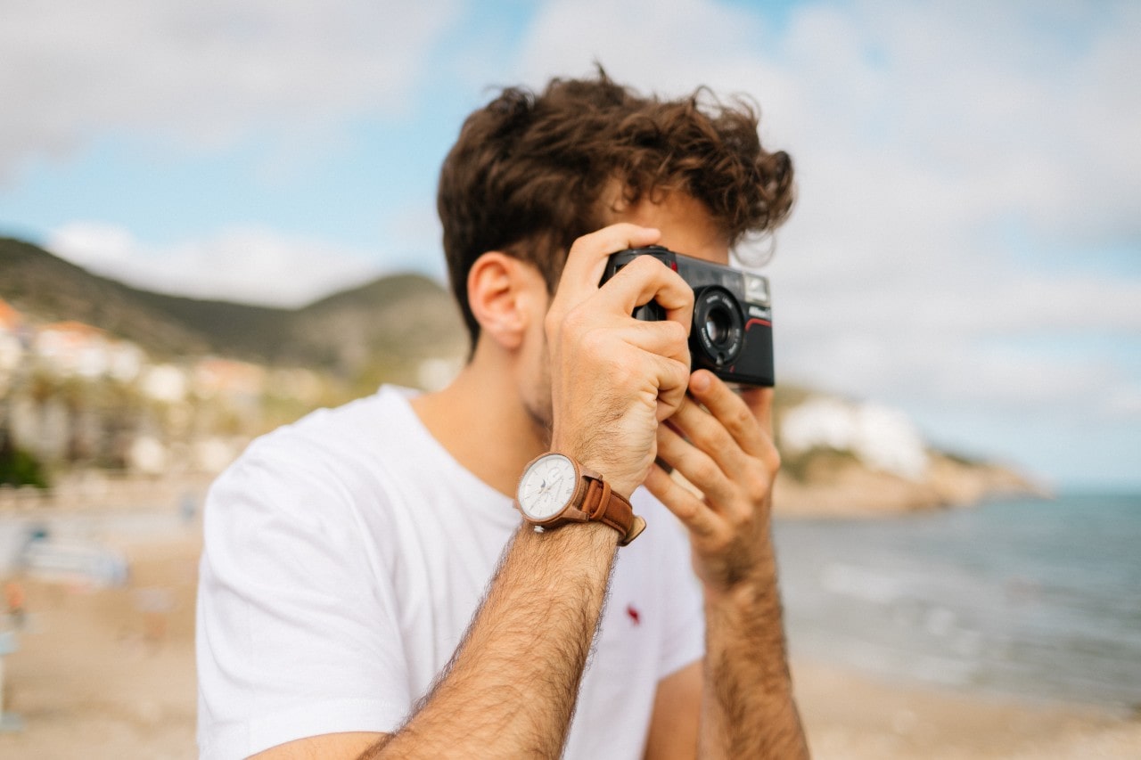 A man on vacation wearing a brown luxury watch while taking a photo of the beach