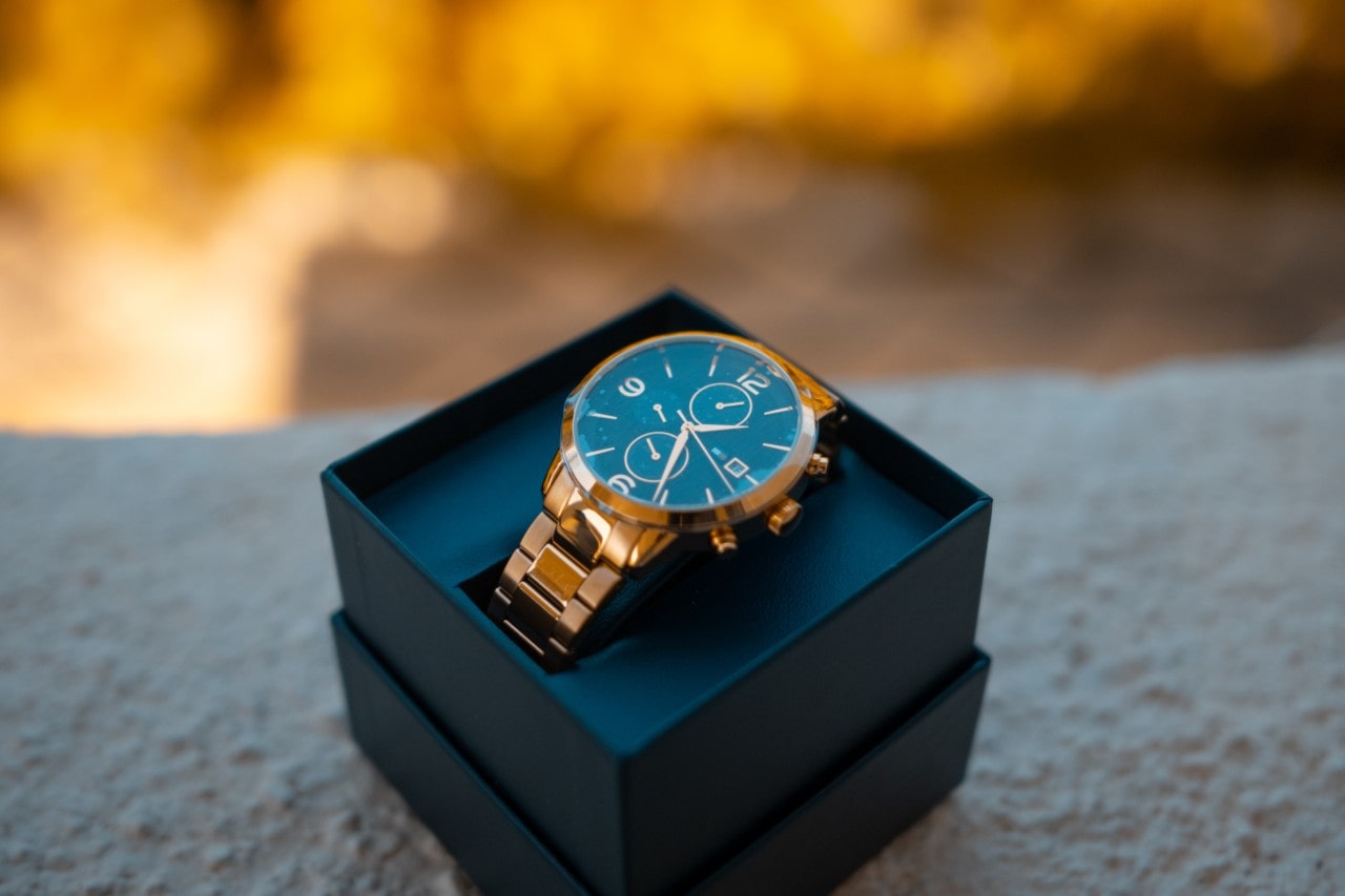 A close-up of a gold watch displayed in-box outside before gold autumn leaves.