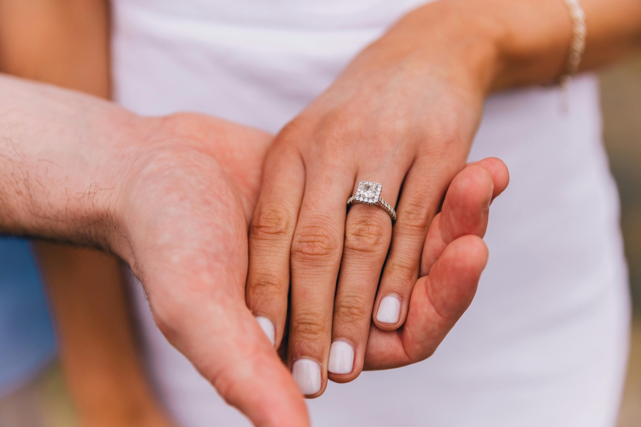 a lady’s hand wearing a diamond engagement ring clasping a man’s hand