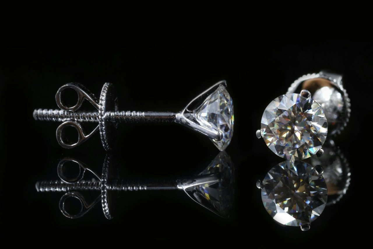 A close-up of a pair of diamond stud earrings on a black backdrop, one displayed from the front and the other from the side. A close-up of a pair of diamond stud earrings on a black backdrop, one displayed from the front and the other from the side.