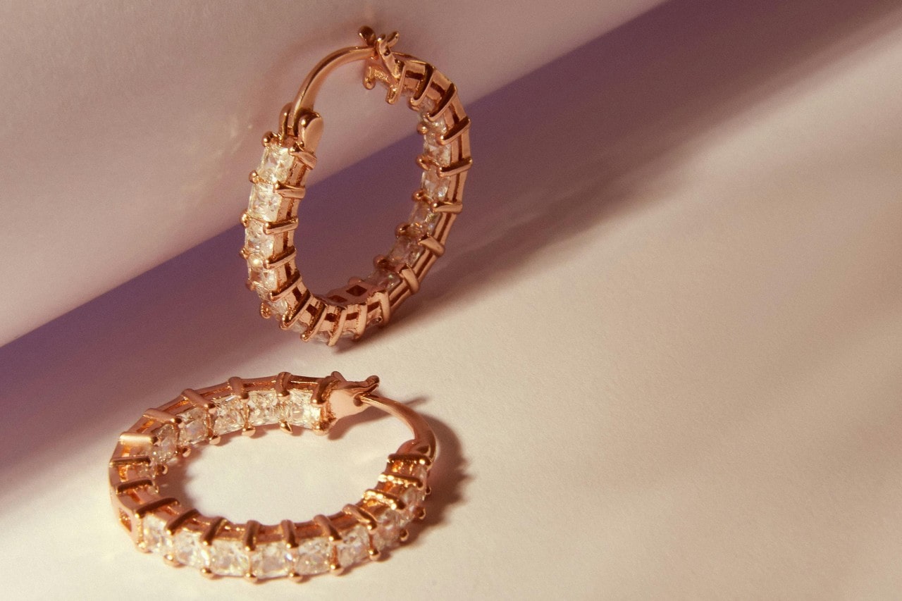 A close-up of a small pair of yellow gold diamond hoop earrings on a light backdrop. A close-up of a small pair of yellow gold diamond hoop earrings on a light backdrop.