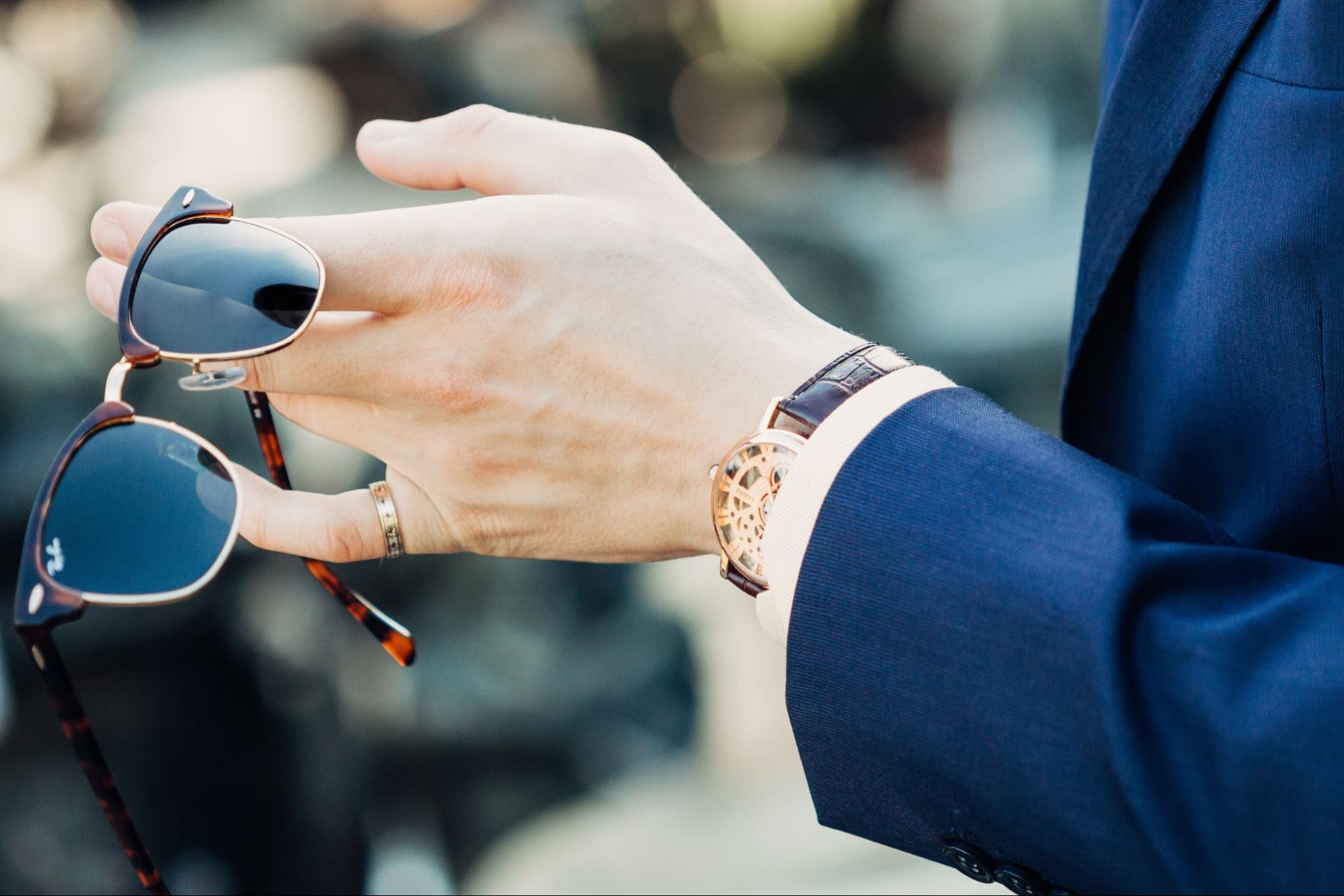 A man in a navy-blue suit holding sunglasses while wearing a luxury skeleton wristwatch with a brown leather strap. A man in a navy-blue suit holding sunglasses while wearing a luxury skeleton wristwatch with a brown leather strap.