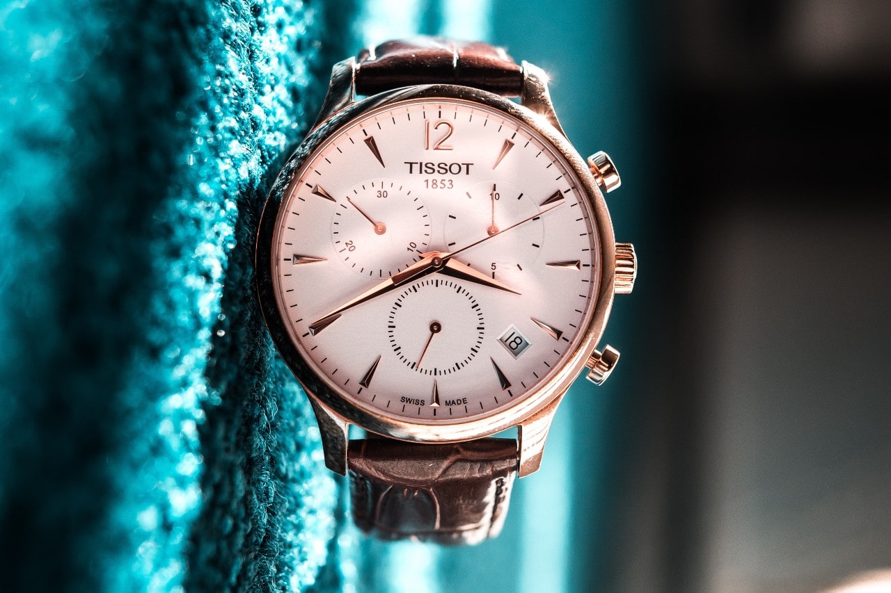 A close-up of a Tissot chronograph wristwatch with a brown leather strap, resting against a textured turquoise fabric. A close-up of a Tissot chronograph wristwatch with a brown leather strap, resting against a textured turquoise fabric.
