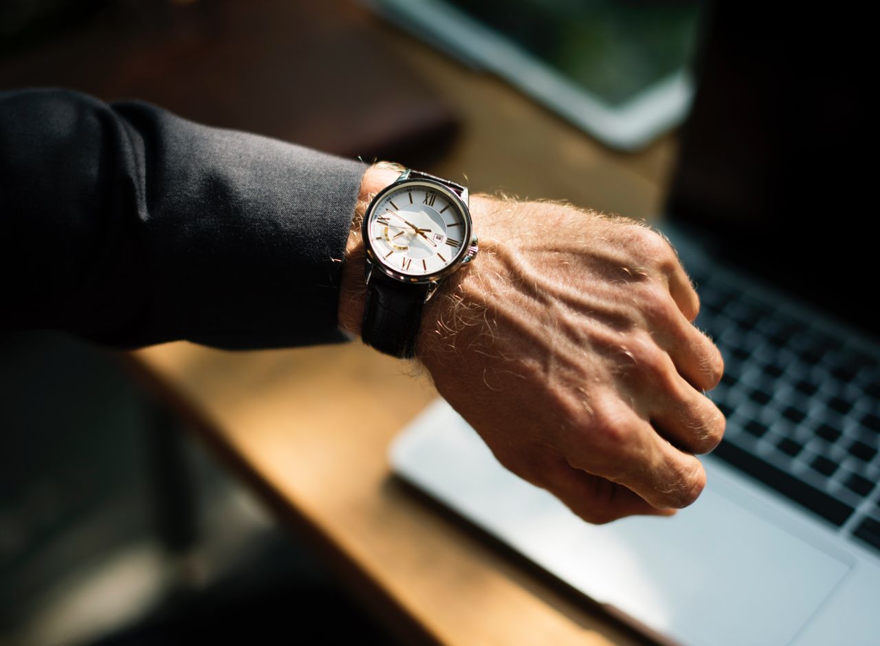 A man’s hand showcasing classic white-dial wristwatch with a black leather against a blurry laptop background A man’s hand showcasing classic white-dial wristwatch with a black leather against a blurry laptop background
