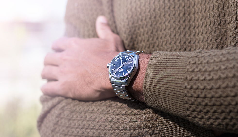 A man wearing a stainless steel wristwatch with a blue dial, crossing his arms over a grey knitted sweater. A man wearing a stainless steel wristwatch with a blue dial, crossing his arms over a grey knitted sweater.