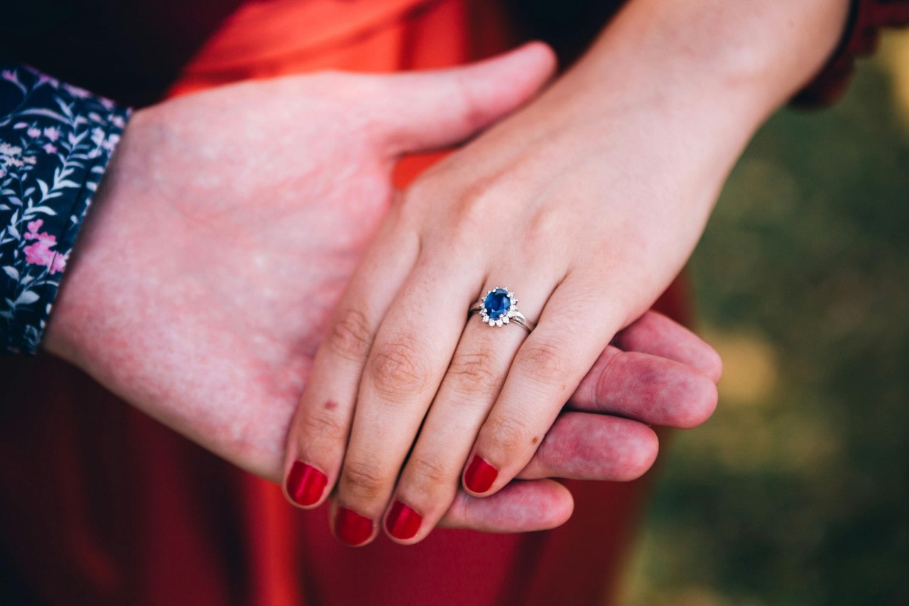 A close-up of a bride-to-be’s hand in her groom’s, her finger adorned with a sapphire engagement ring.