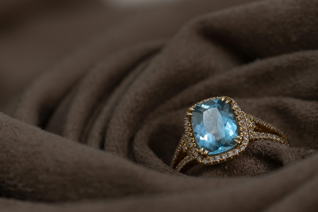 A close-up of an aquamarine halo engagement ring nestled within a blanket.