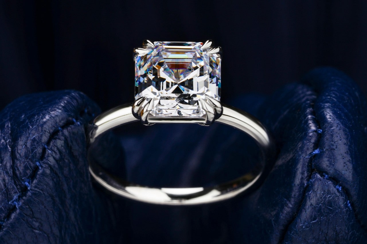 A close-up image of an asscher cut diamond ring held between gloved fingers.