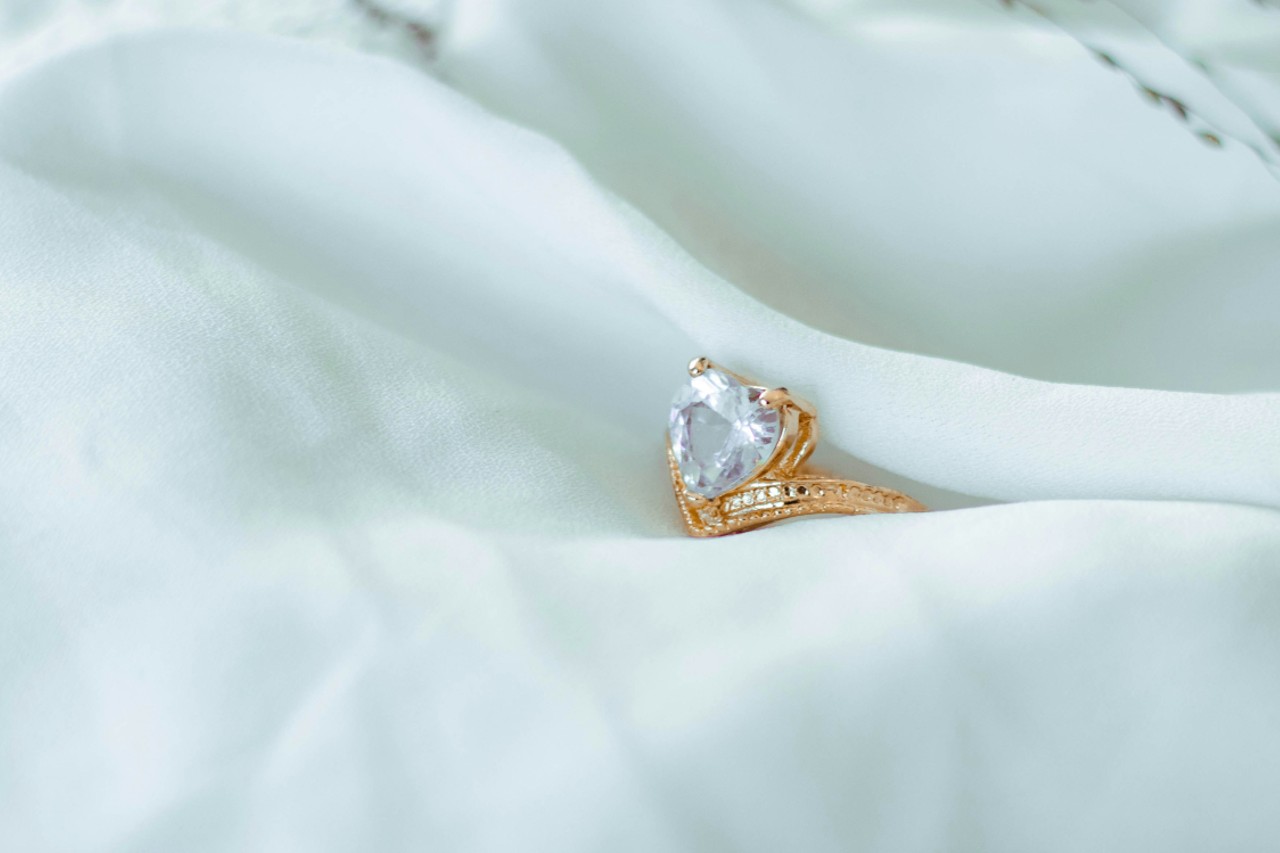 A yellow gold diamond ring with a heart shaped center stone, nestled in pale fabric.