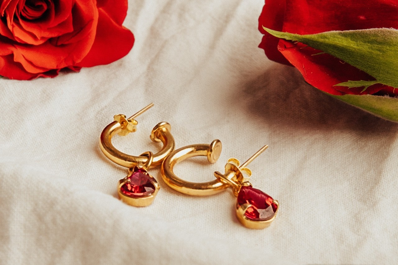 a pair of gold earrings with red gemstones on a table cloth next to flowers