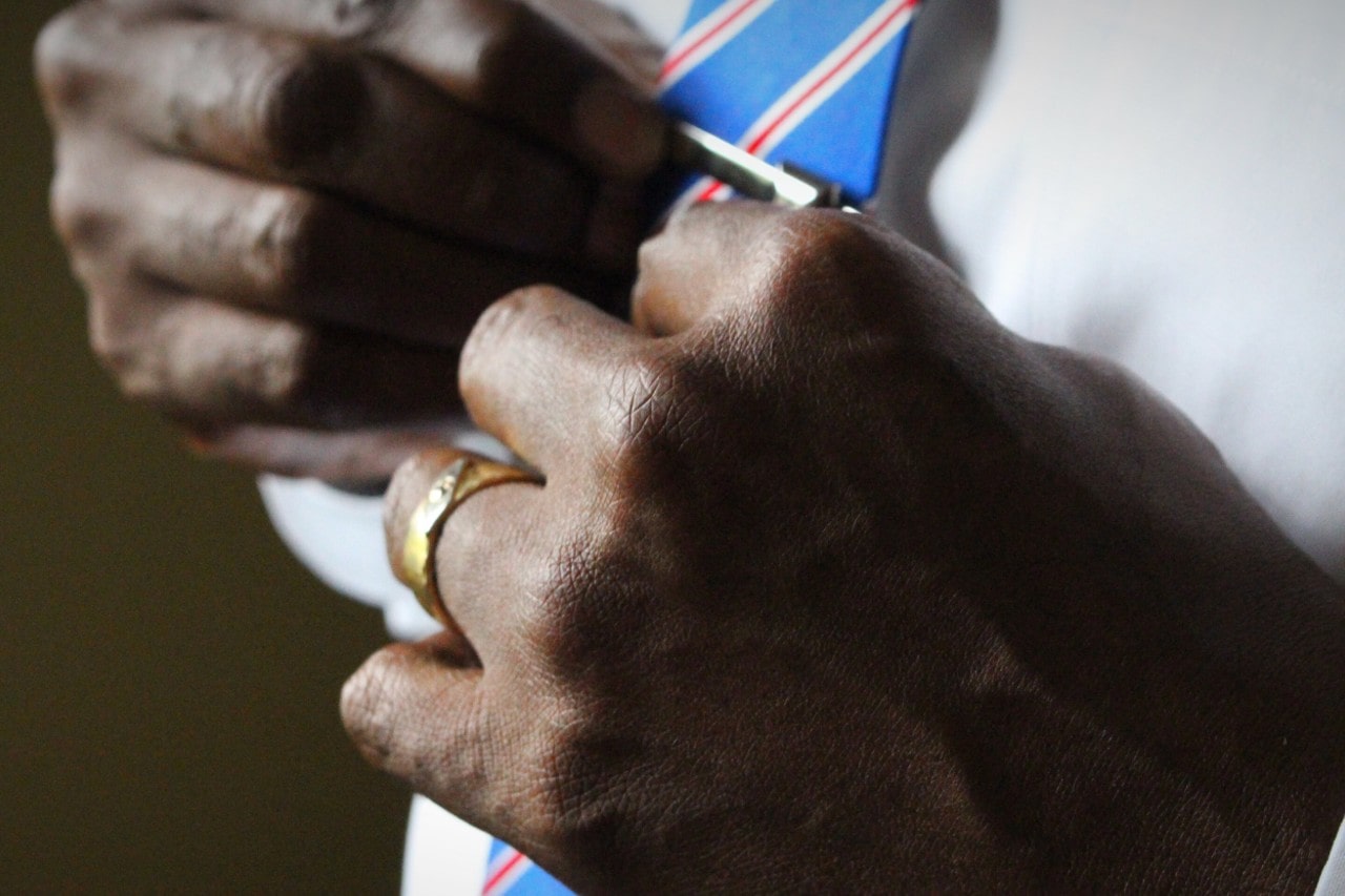 A close-up image of a man adjusting his tie clip, wearing a mixed metal engagement ring.