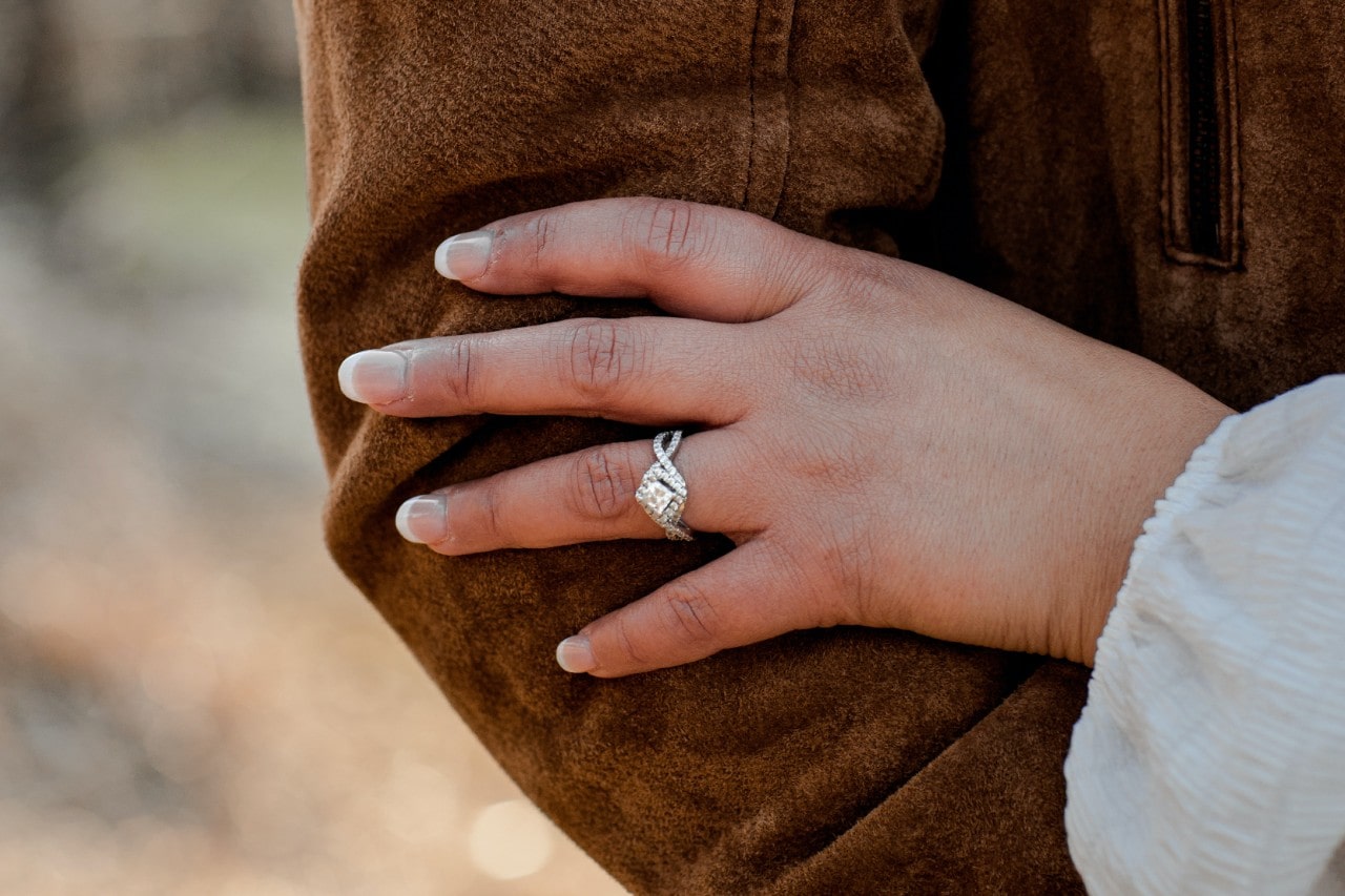 A woman wearing a halo ring lays her hand on a man’s leather jacket sleeve.