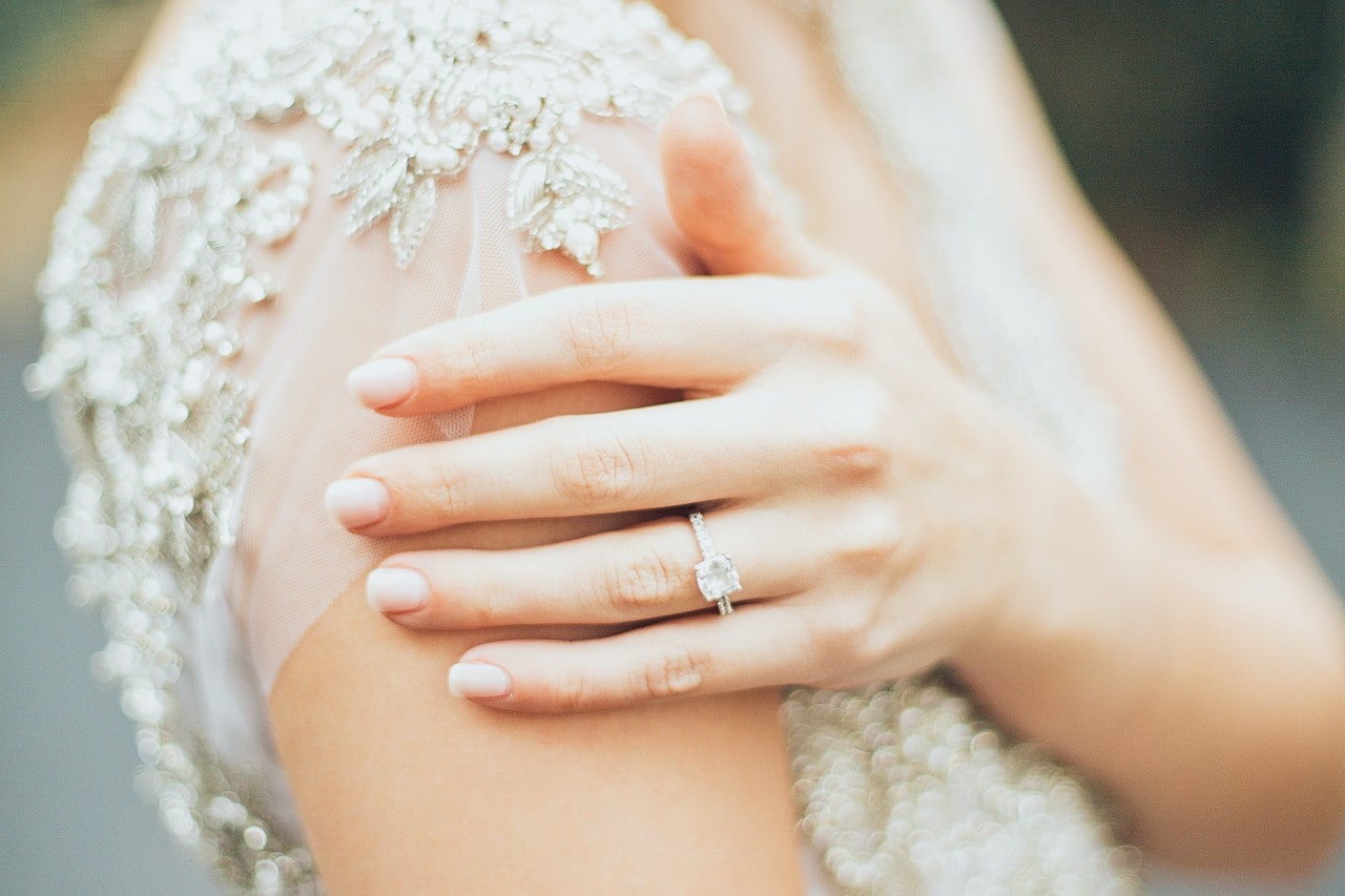 A bride rests her hand on her shoulder, wearing a princess cut ring.